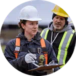 Two HVAC professionals in hard hats and safety vests have a discussion. One holds a clipboard, while the other looks on.