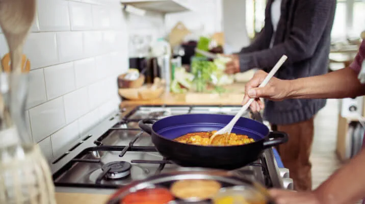 Close-up view of an adult using a wooden spoon to stir a skillet. Another stands in the background while chopping vegetables.