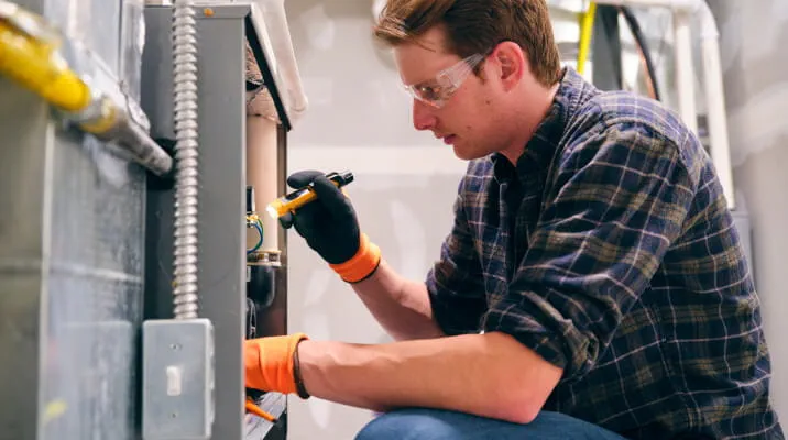 A contractor wearing safety goggles and gloves uses a flashlight to examine an HVAC system.