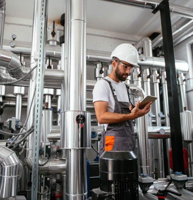 Man wearing hard hat and protective goggles looking at tablet in boiler room