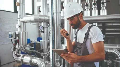 Engineer holds tablet and walkie-talkie in boiler room.