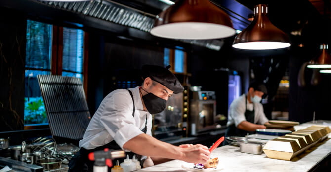 Two male chefs wearing protective face masks and preparing food in kitchen