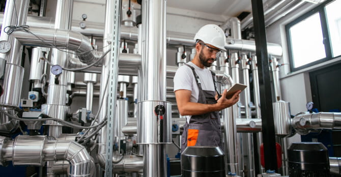 Man wearing hard hat and protective goggles looking at tablet in boiler room