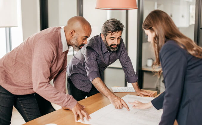 Three business colleagues examining and discussing a blueprint while standing at a boardroom table. 