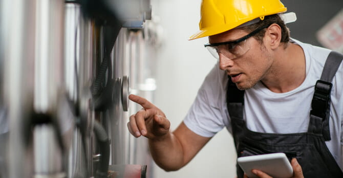 Man in hard hat and safety goggles inspecting natural gas processing machine and holding tablet