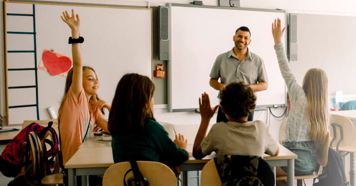 A group of 4 schoolchildren sit around a desk in a modern classroom. Two students (one to the right side of the table and the other to the left side) eagerly raise their hands, while one in the middle partially raises their hand. A teacher stands at the front of the room, smiling.