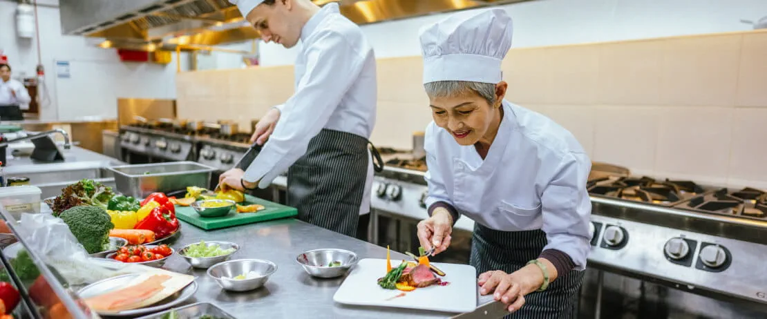 Two cooks work in a well-lit kitchen space, preparing meals using a colourful array of vegetables.