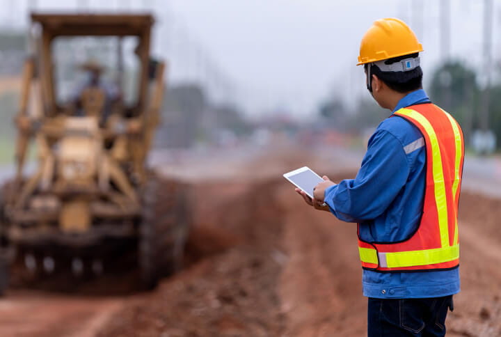 A contractor in safety gear overseeing a road grader on a construction site.