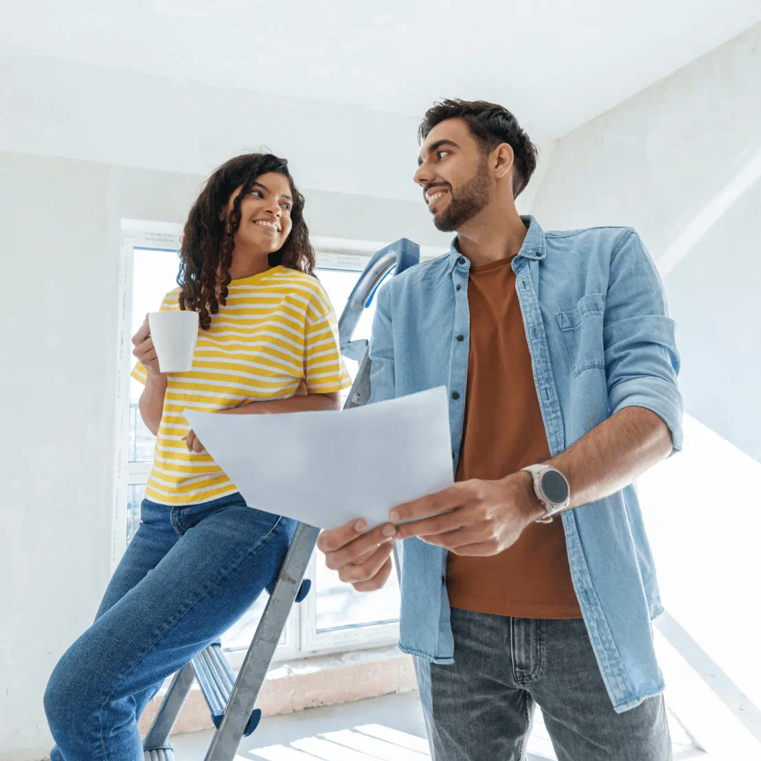 A happy couple smile at each other. One showcases a blueprint sheet, while the other holds a coffee mug.