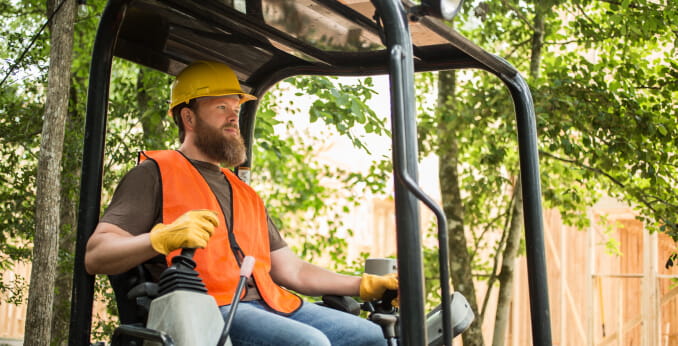 A contractor in a safety vest and hardhat operating a small excavator in a backyard.